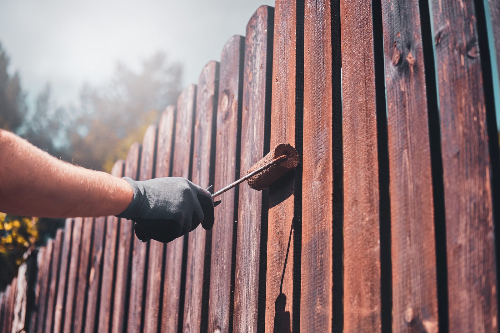 Worker in gloves painting and renovating a wooden fence on a sunny day.